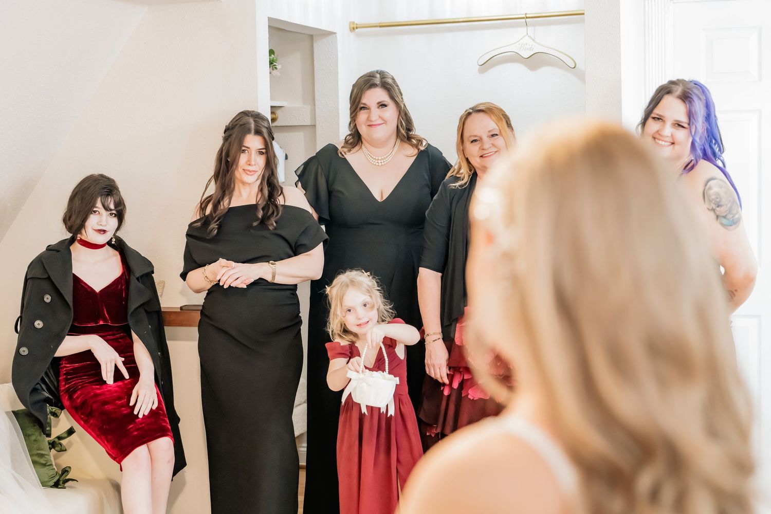 Bridesmaids in black dresses and a flower girl in red watch the bride during wedding preparations.