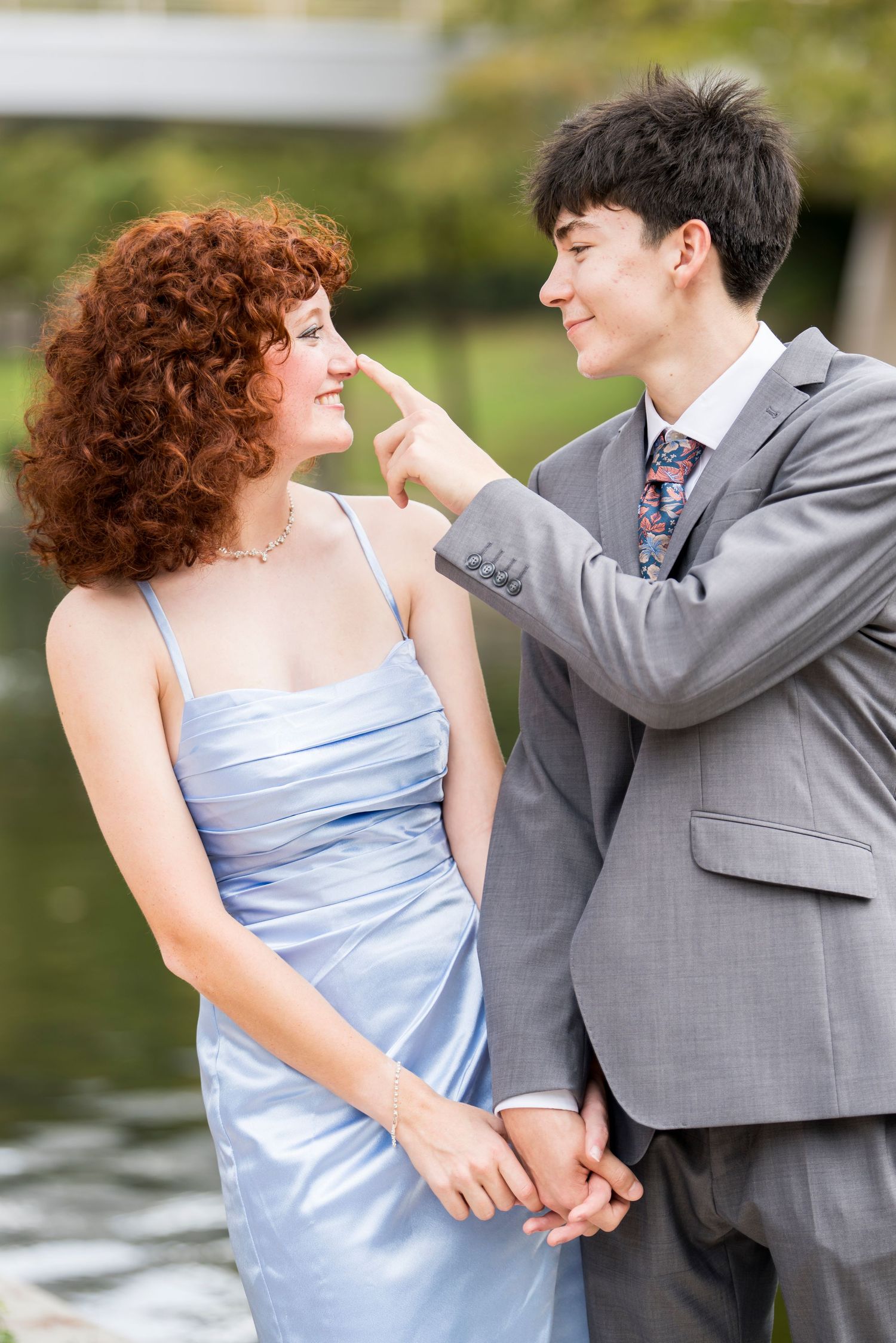 Young couple in formal wear smiling at each other outdoors, man in gray suit touching woman's nose.