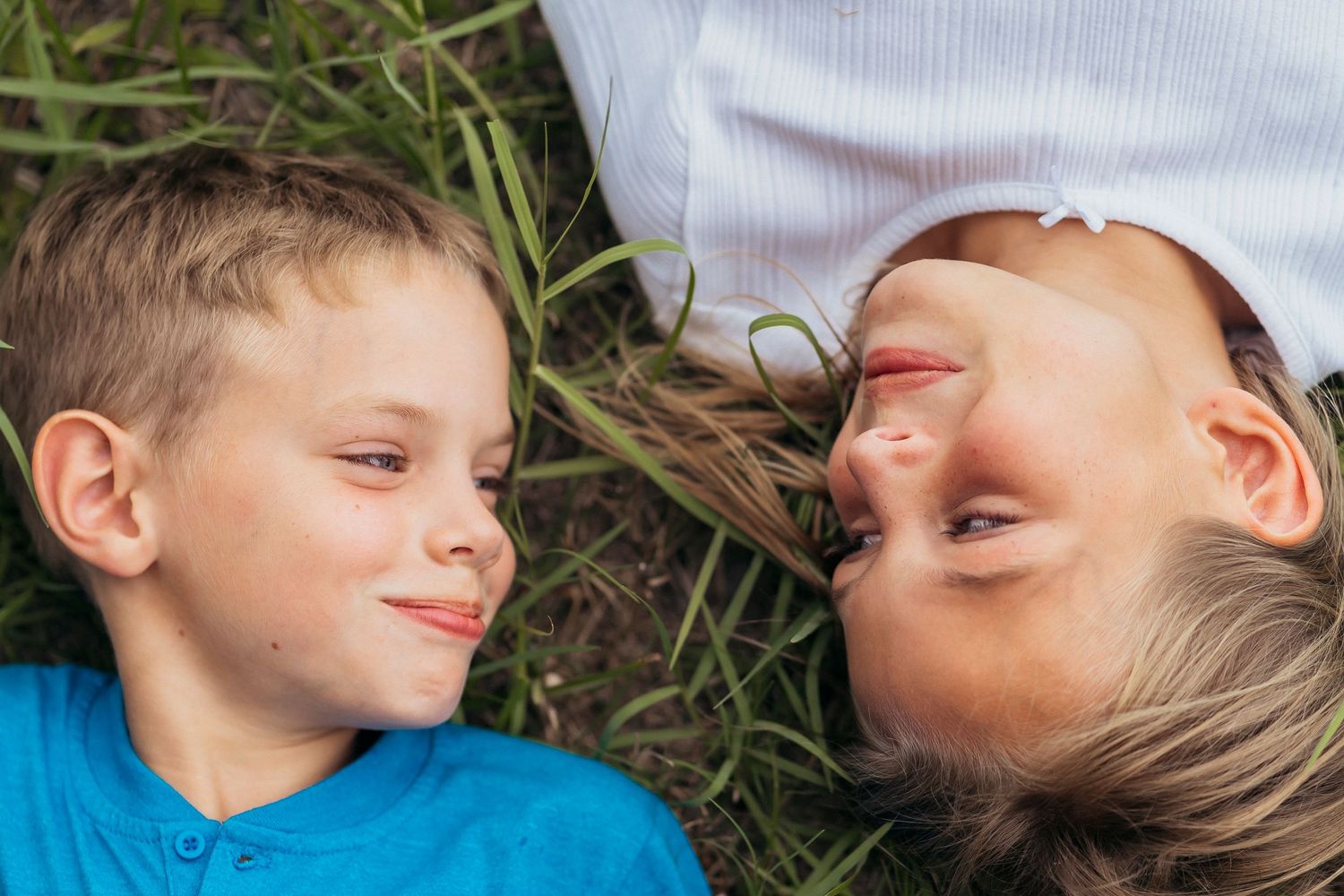 Two happy children lying on green grass, smiling at each other on a sunny day.