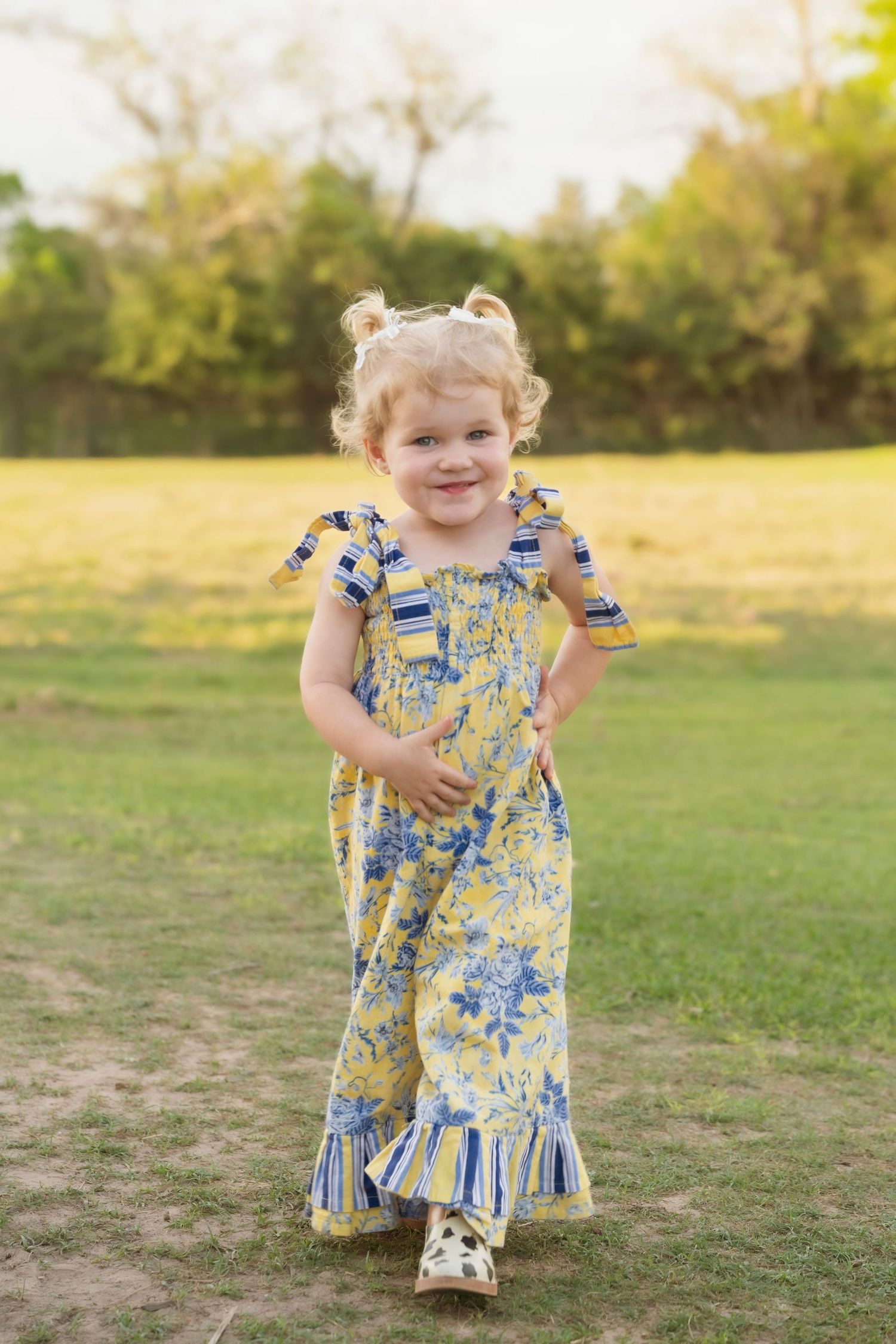 Smiling toddler girl in yellow floral jumpsuit running on green grass in a sunny outdoor park setting.