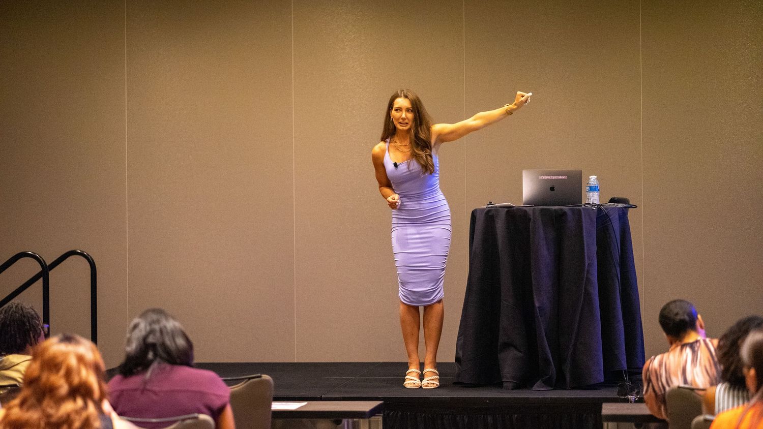 Woman in lavender dress presenting at a conference, pointing toward a screen in a modern event hall.
