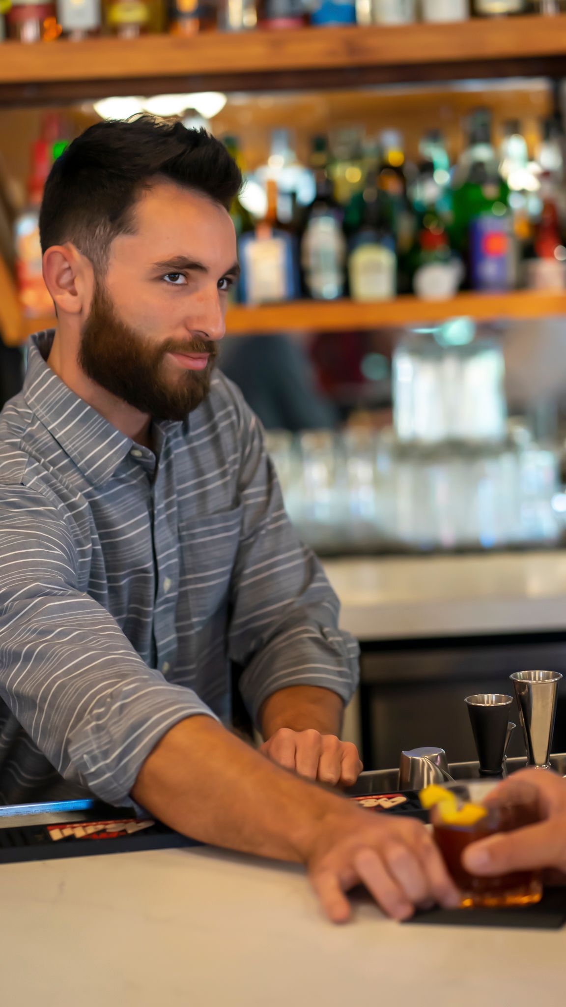 Bearded male bartender serving cocktail drinks behind a well-stocked bar counter.