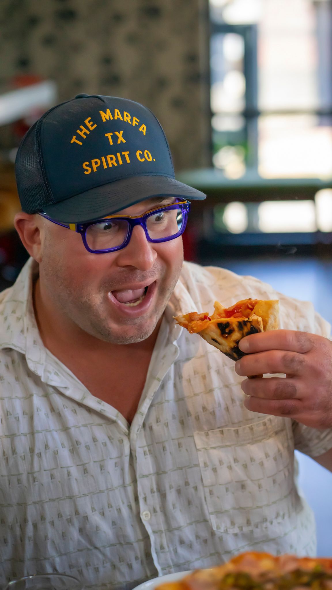 A man wearing a Marfa TX Spirit Co. cap and blue glasses enthusiastically eating a slice of pizza.