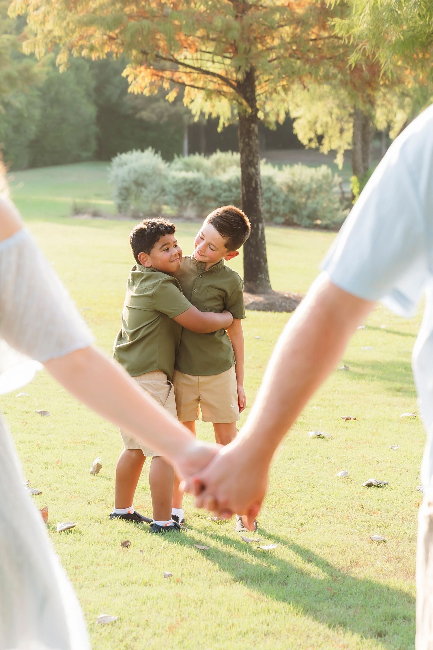 Two boys in matching green shirts hugging in a sunny park while adults hold hands in the foreground.
