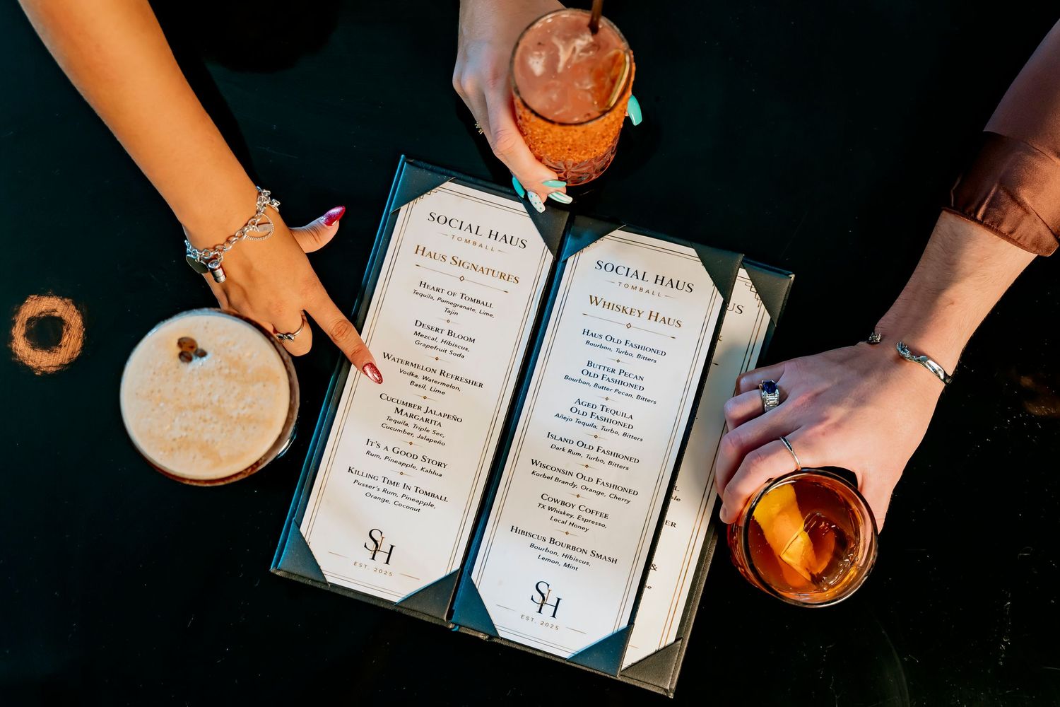 Two people holding cocktails over a Social Hour bar menu on a dark table with drinks and snacks.
