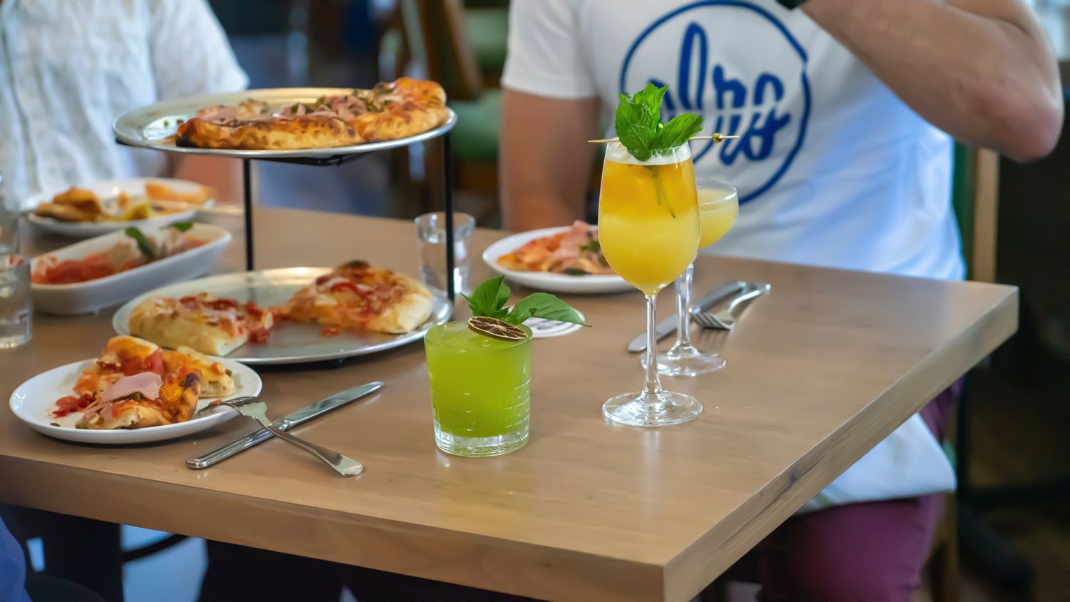 A table set with colorful cocktails, green juice, and pizza at a casual restaurant dining setting.