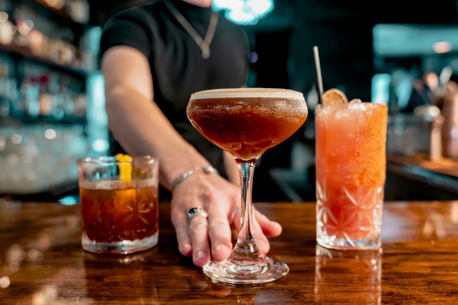 Bartender presenting three colorful cocktails on a wooden bar, including a espresso martini and two red drinks.