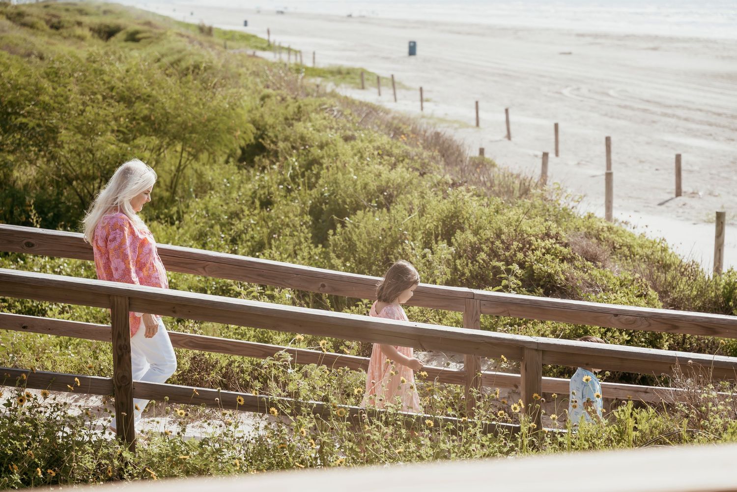 Grandmother and grandchildren walking on a wooden boardwalk path near a sandy beach on a sunny day.
