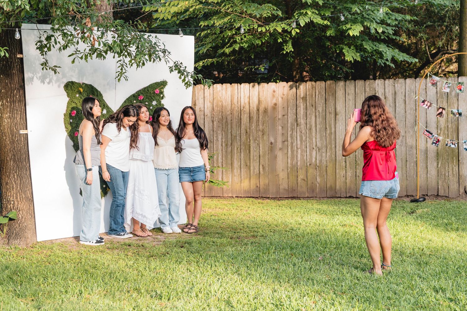Girls posing in front of a white backdrop with a butterfly mural in a backyard while a friend takes their photo.