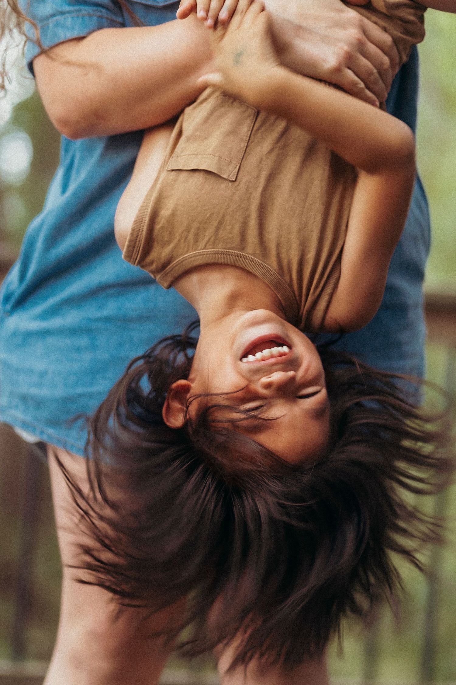 A laughing child held upside down by an adult outdoors, with dark hair flowing and wearing a tan shirt.