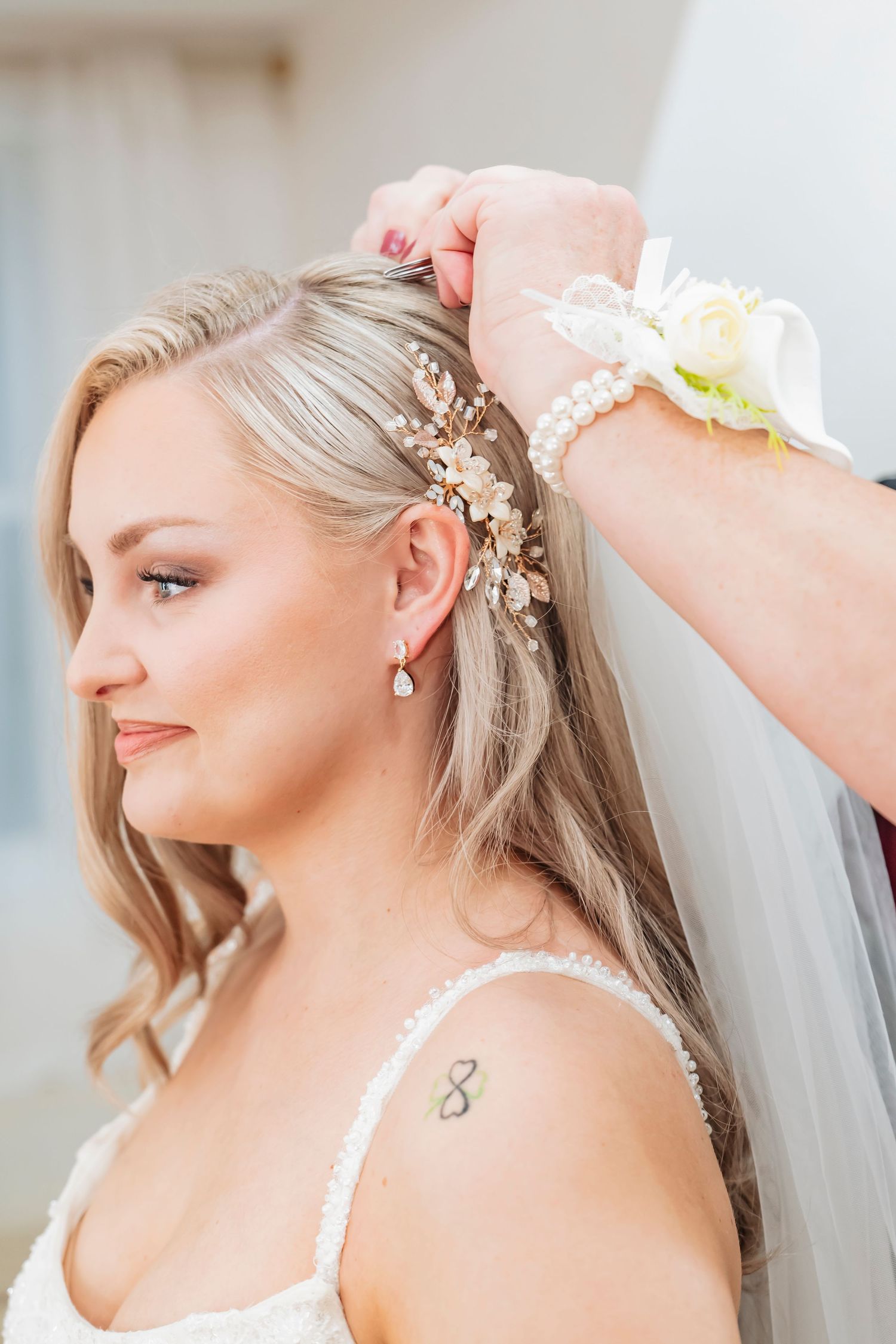 Bride getting hair accessory placed by assistant wearing floral wrist corsage, with veil, pearl earrings, and infinity tattoo.