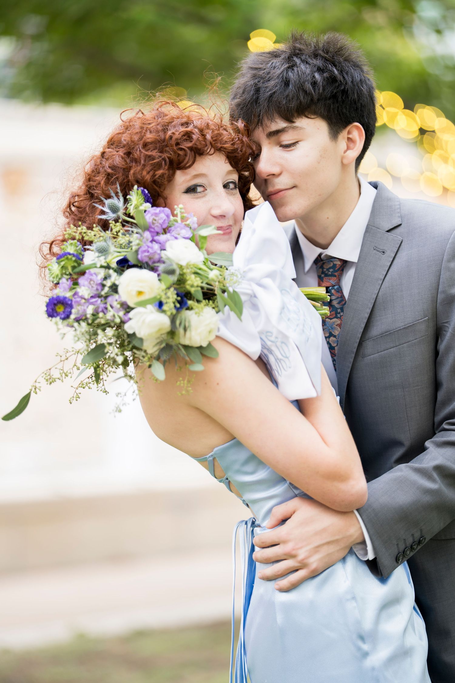 A groom in a gray suit embraces a bride with red curly hair holding a purple and white bouquet outdoors.
