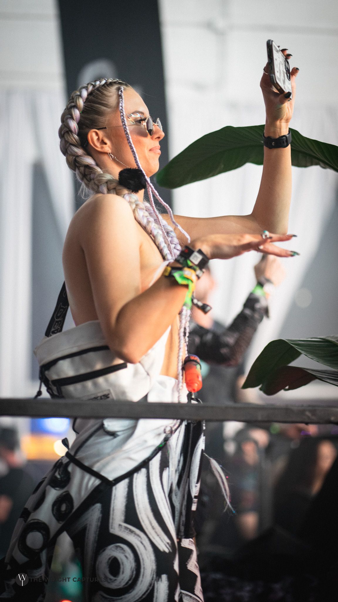 Woman with braided hair takes selfie at festival, wearing bold black and white outfit with colorful accessories.
