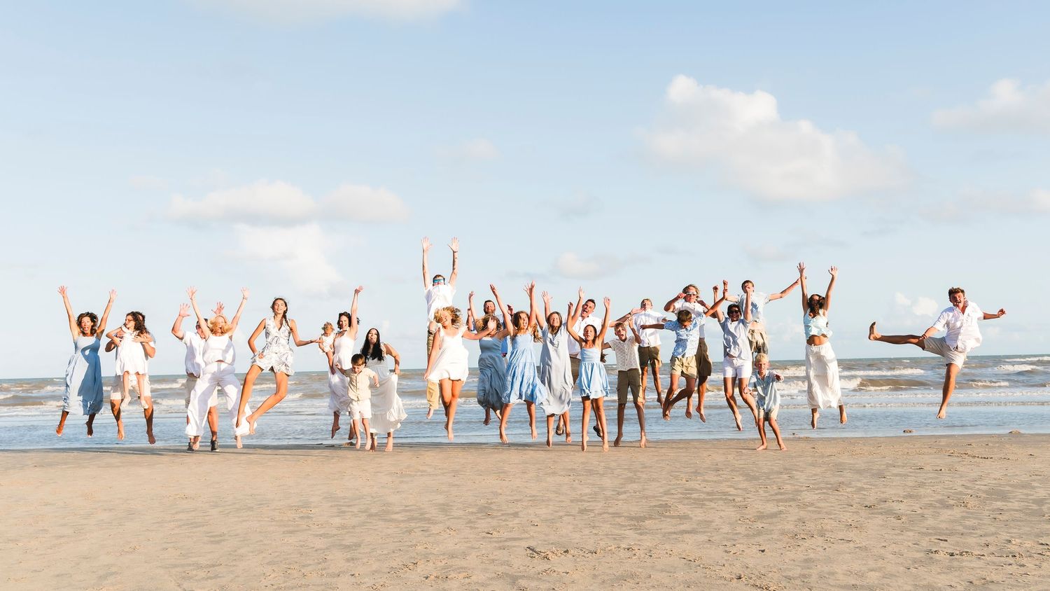 Large group of joyful people jumping on a sunny beach, all dressed in white and light colors.