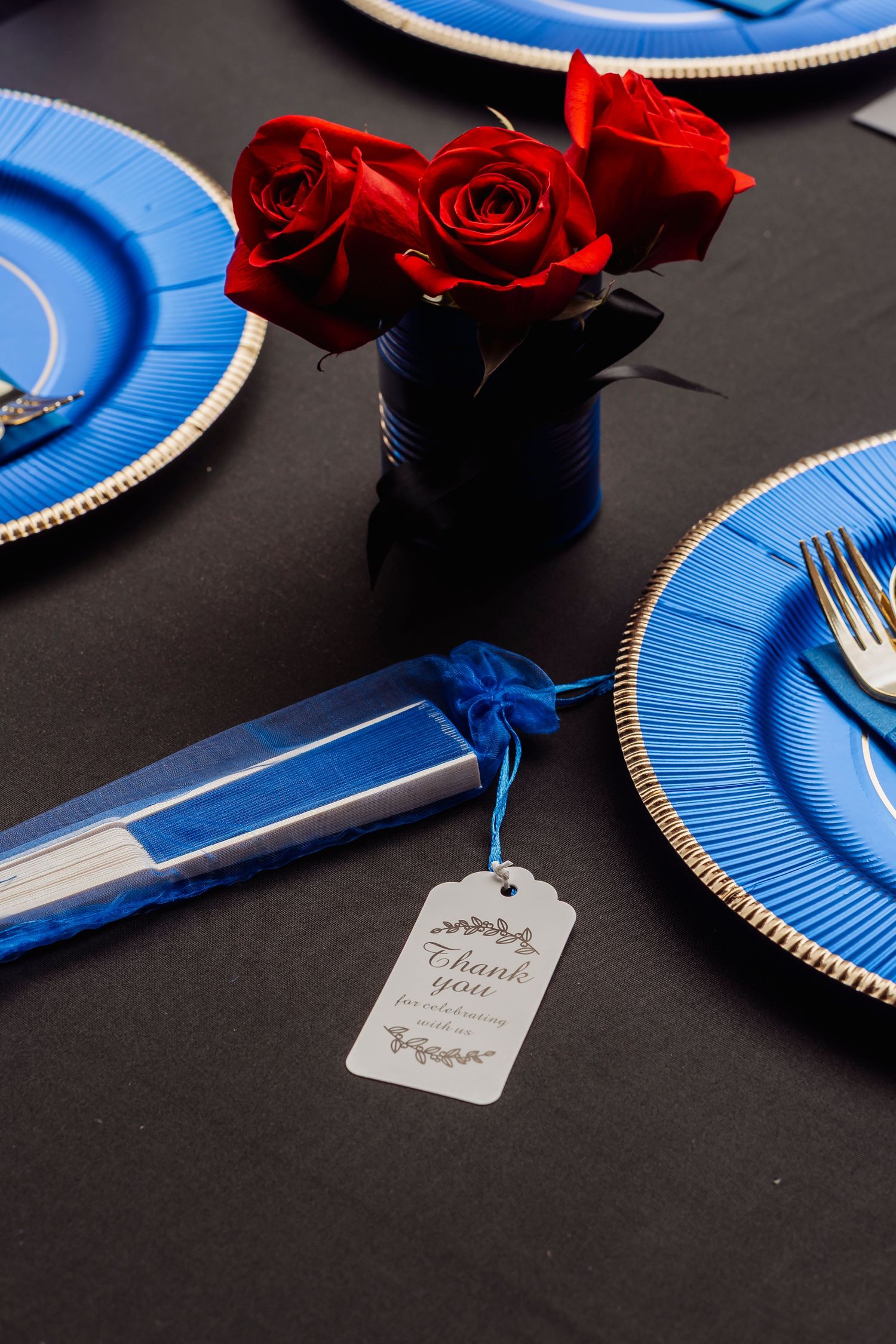 Elegant table setting with blue charger plates, gold cutlery, red roses centerpiece, and thank you gift tag on black tablecloth.