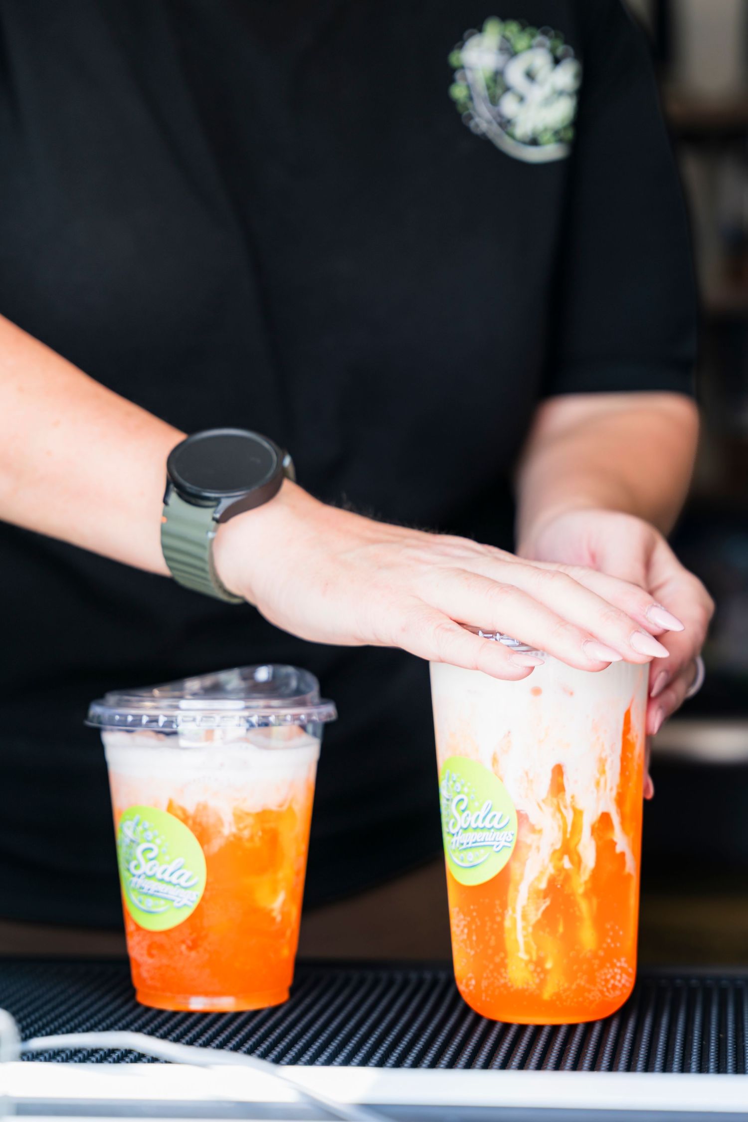 Barista preparing two vibrant orange boba drinks with cream tops in branded cups at a cafe counter.