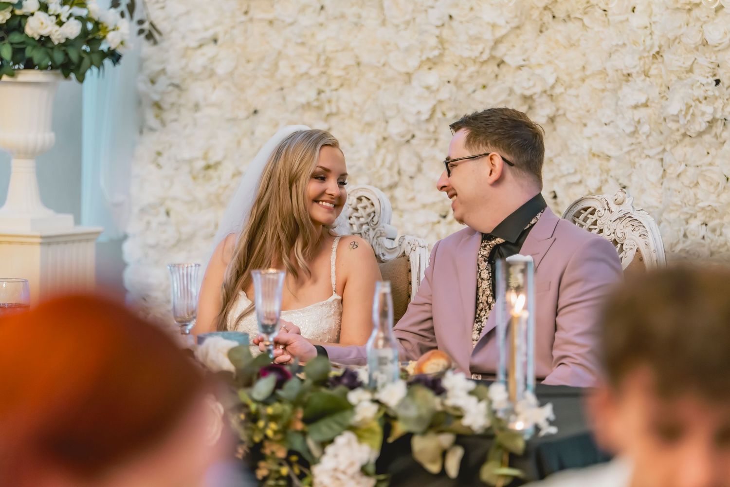 Bride and groom smiling at each other at wedding reception table with floral wall backdrop.