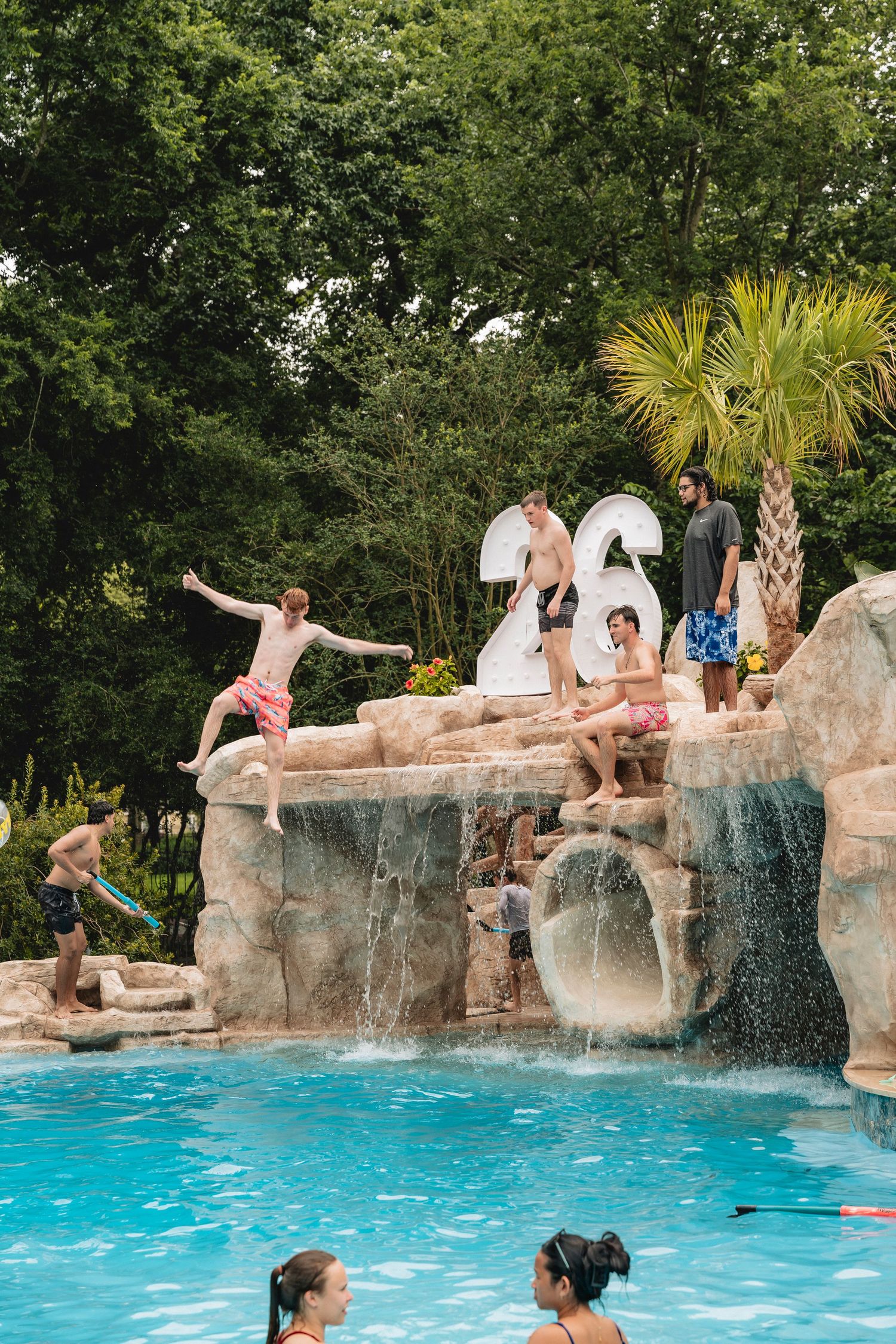 Kids jumping off rocky waterfall feature into a resort-style backyard pool surrounded by lush trees.