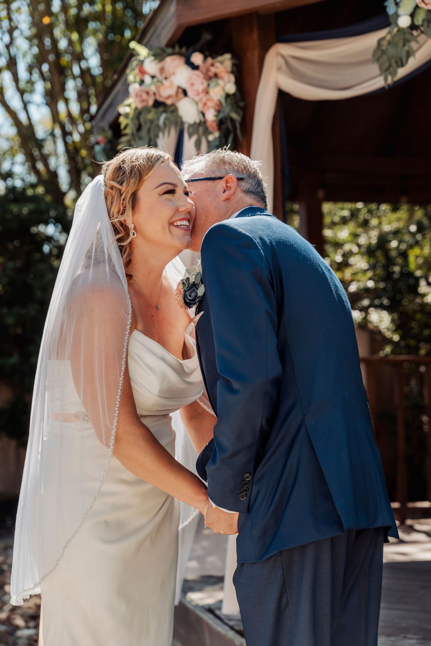 A bride in a white gown and veil shares a tender kiss with a man in a navy suit at an outdoor wedding ceremony.