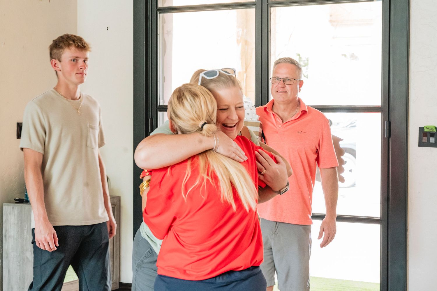 Two women in coral tops share a warm hug near a doorway while a man and teenage boy look on smiling.