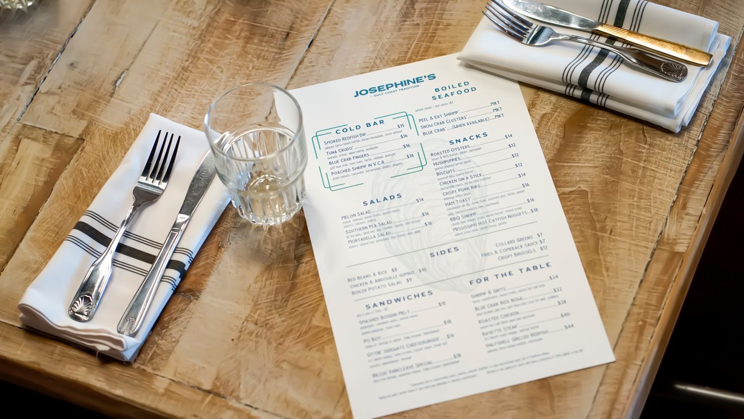 Restaurant menu on wooden table with silverware, napkin, and water glass place setting.