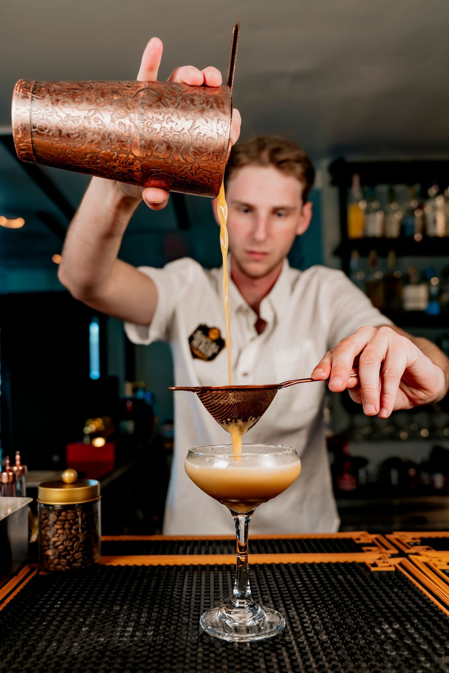 Bartender pouring cocktail through strainer into coupe glass using copper shaker at bar.