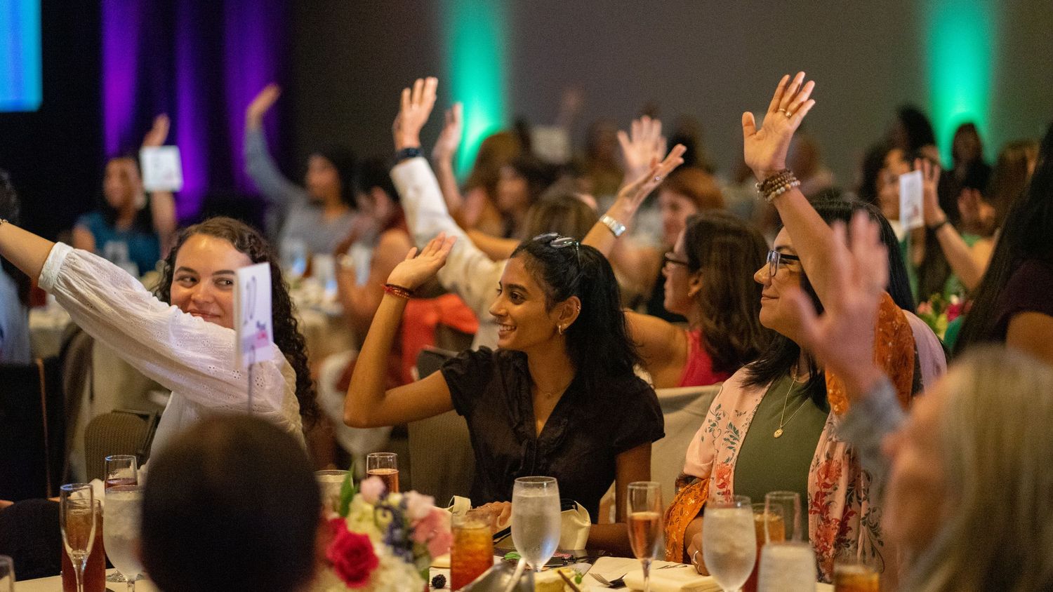 Women raising hands and smiling at a festive banquet event in a decorated conference hall.
