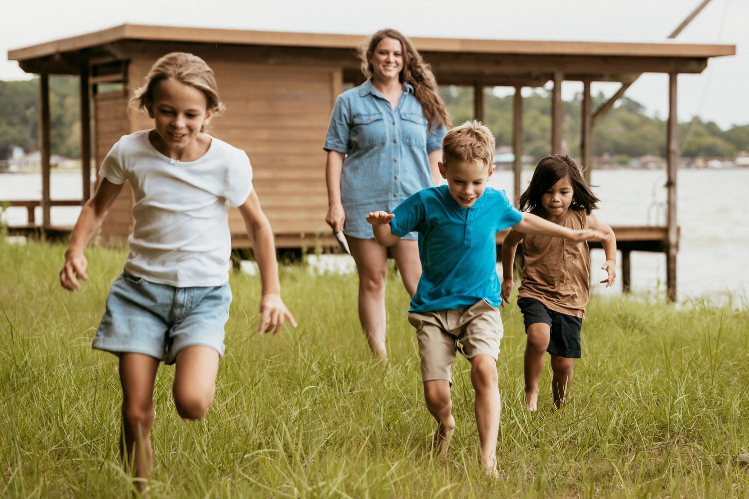 A woman watches three children running through green grass near a lakeside cabin on a sunny day.