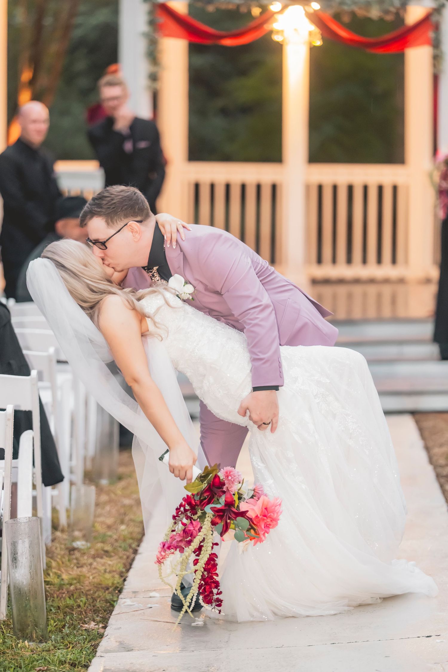 Groom in lavender suit dips bride in white gown for a romantic kiss at outdoor wedding ceremony.