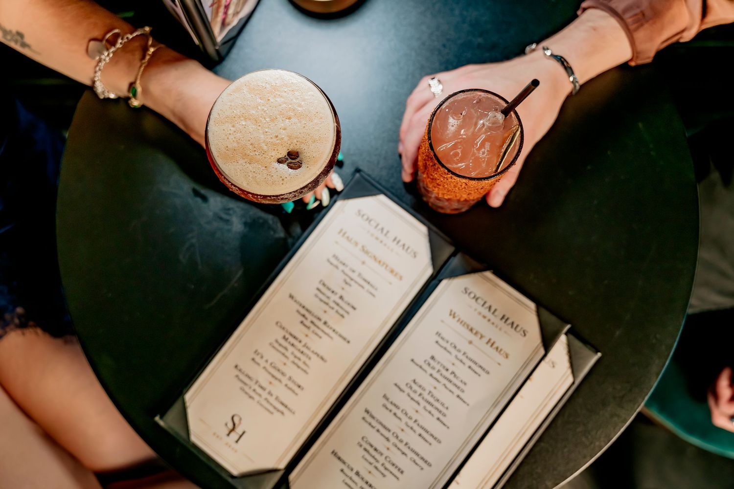 Two cocktails held over open menus on a dark round table at a trendy bar or restaurant.