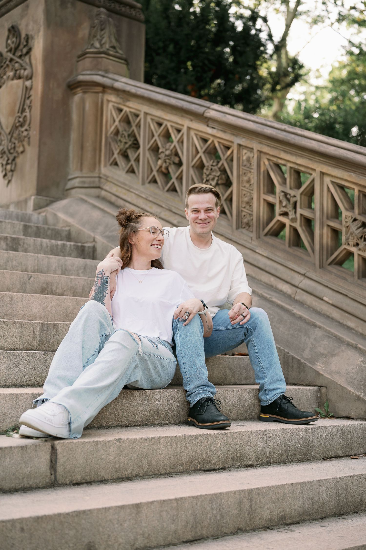 Couple sitting on ornate stone steps, woman with tattoos in white tee and jeans, man in cream sweater, smiling together.