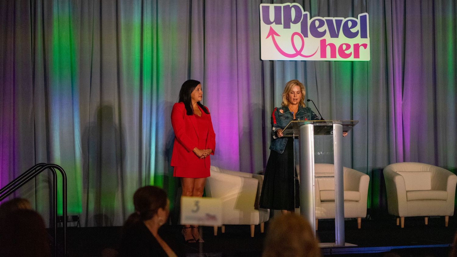 Two women speaking on stage at Up Level Her conference, one at podium, with colorful lighting backdrop.