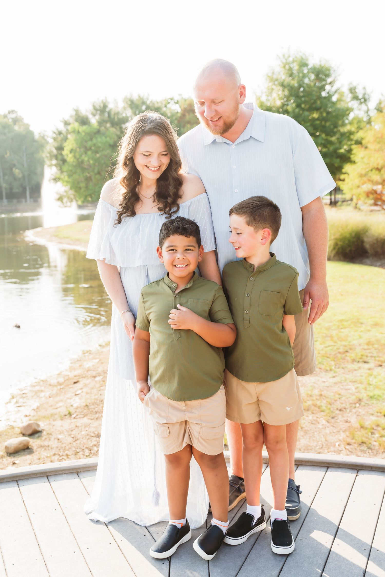 Happy family of four posing outdoors near a pond on a sunny day, wearing coordinated casual outfits.
