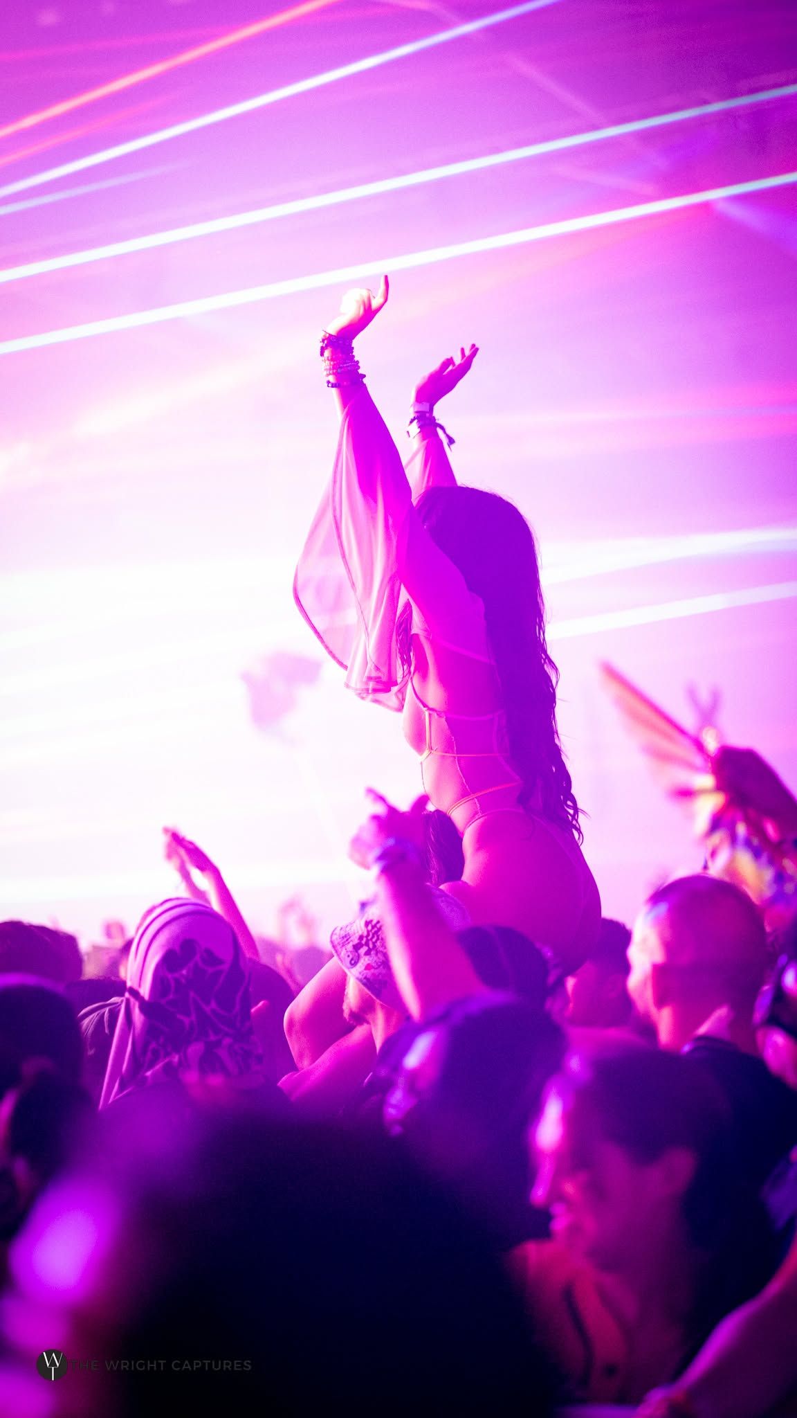 A woman dances elevated above a crowd at a nightclub, bathed in vibrant pink and purple laser lights.