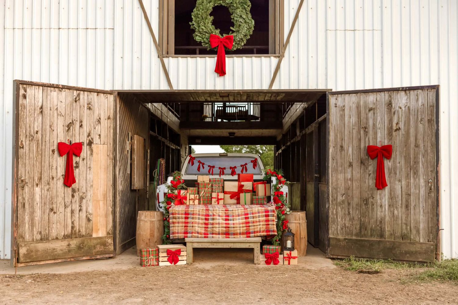 Rustic barn decorated for Christmas with wreaths, red bows, and wrapped gifts on a plaid bench.