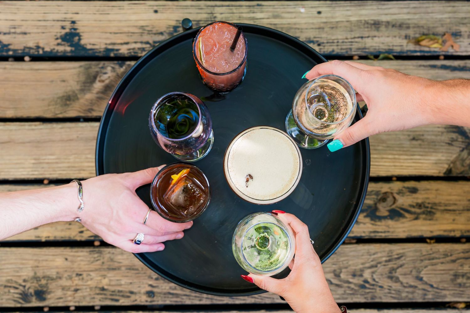 Overhead view of friends toasting with various drinks on a round black tray on a wooden table.