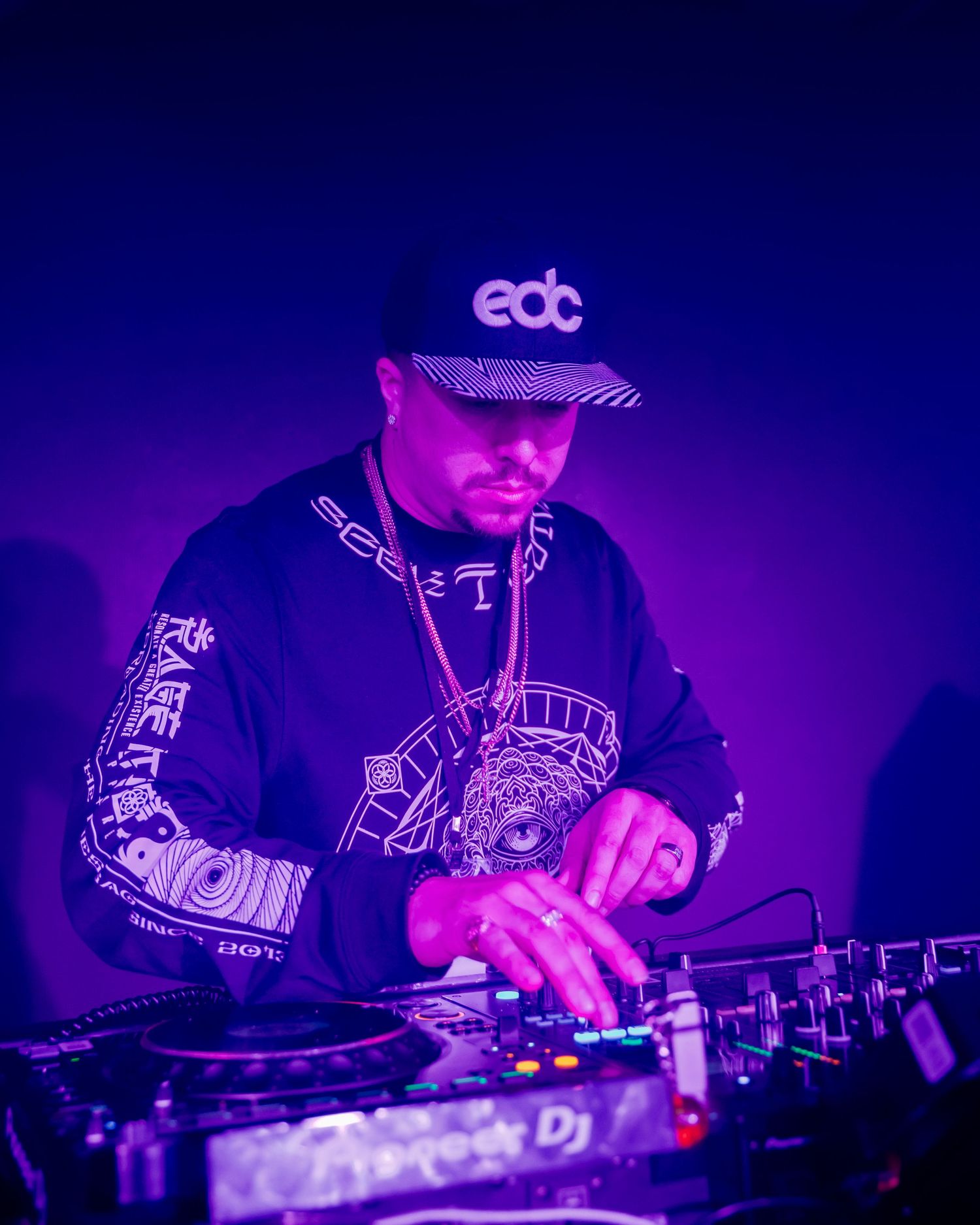 A DJ wearing an EDC cap performs under purple lighting at a music event, mixing tracks on professional turntables.