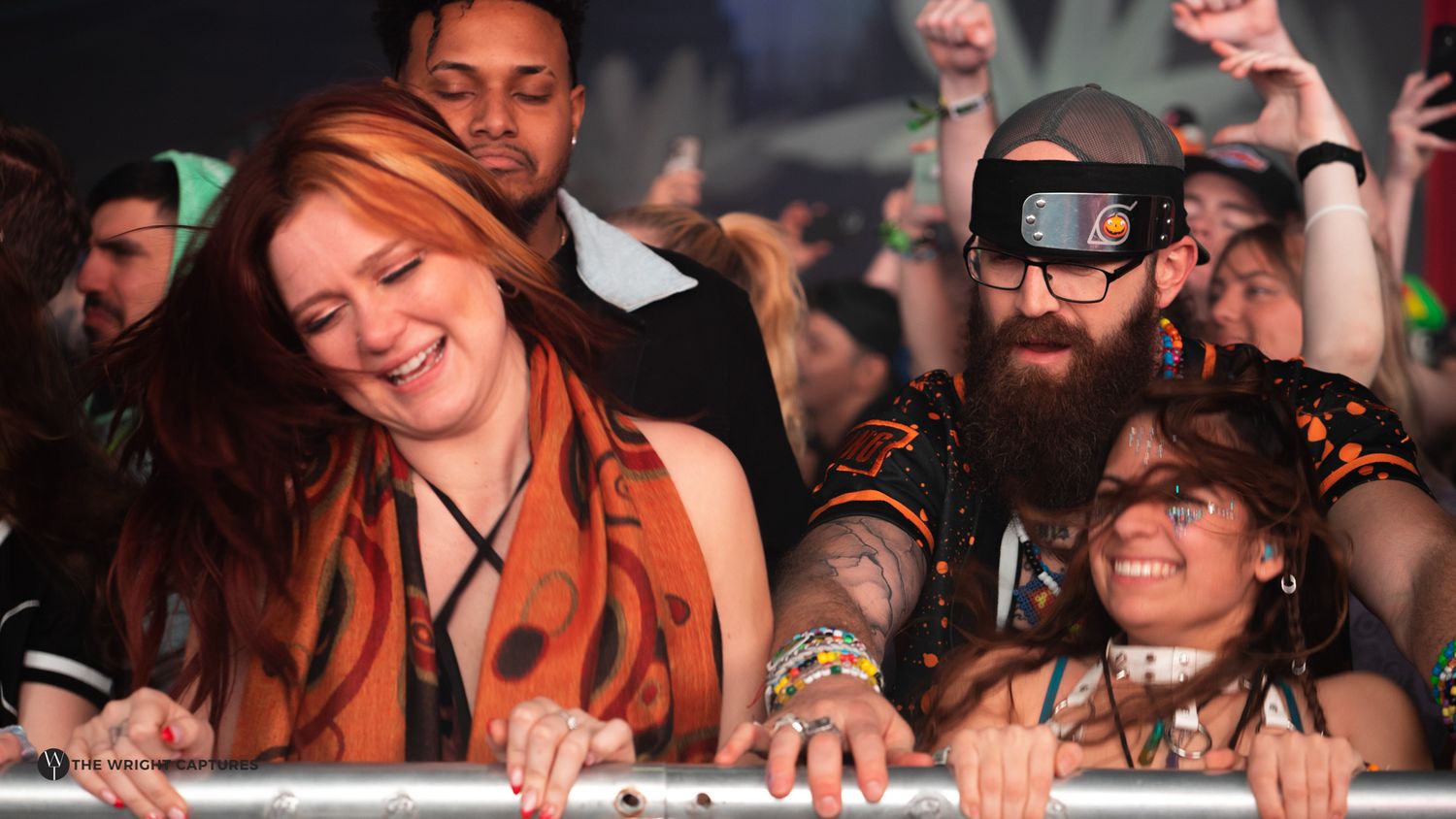 Crowd of festival-goers smiling and leaning on a barrier, including a bearded man wearing futuristic goggles.