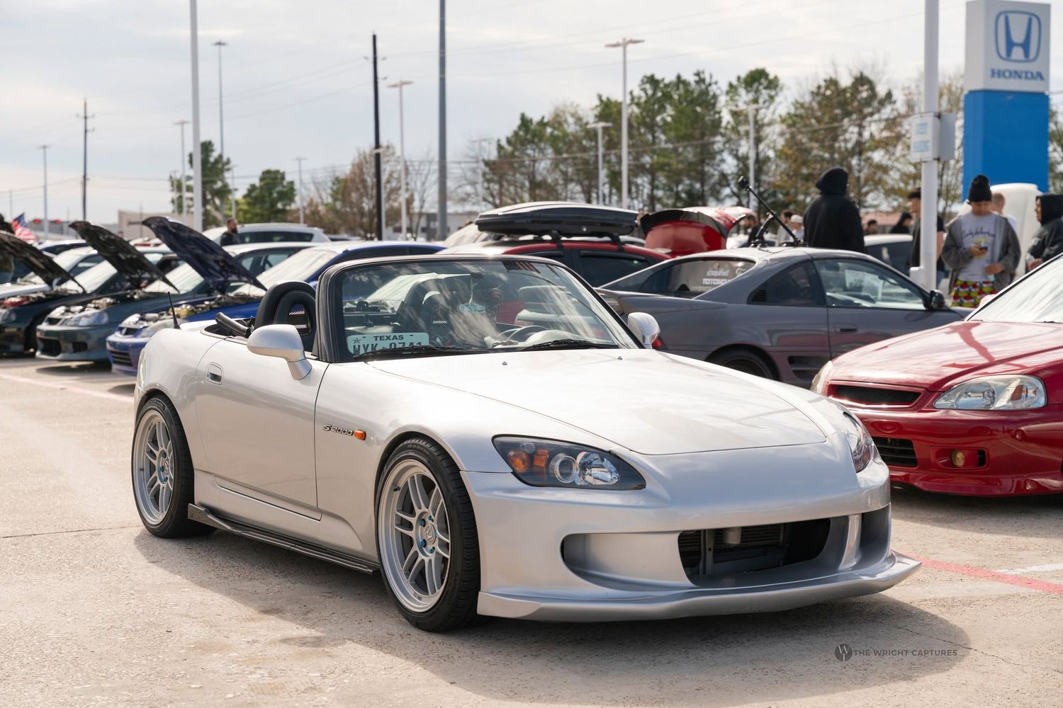 White Honda S2000 sports car displayed at an outdoor car show with other vehicles and a Honda dealership in the background.