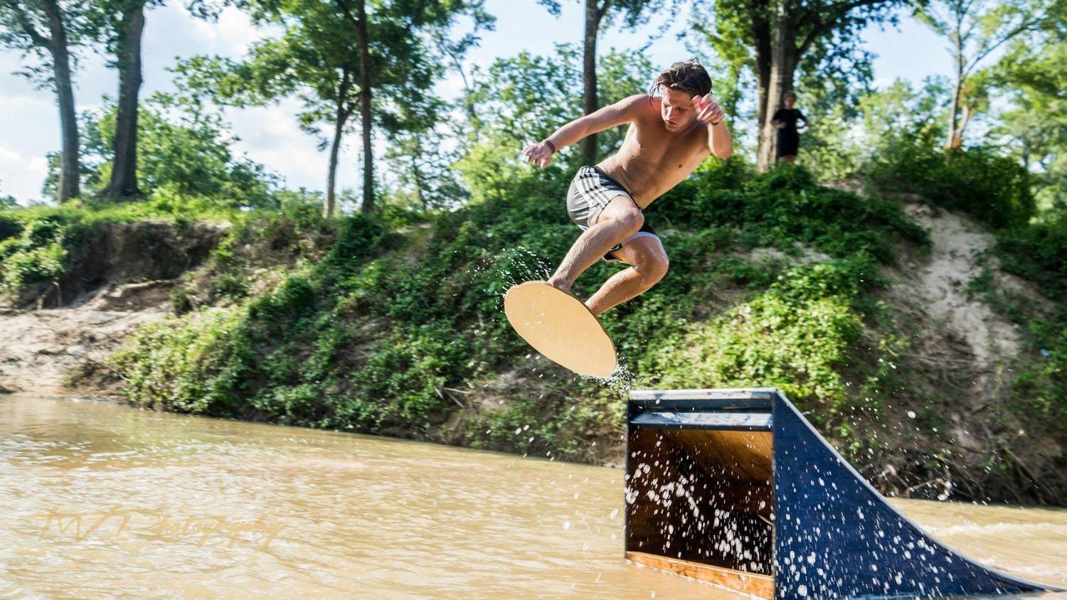 Shirtless man performing a wakeskate trick off a metal ramp into muddy brown water outdoors.