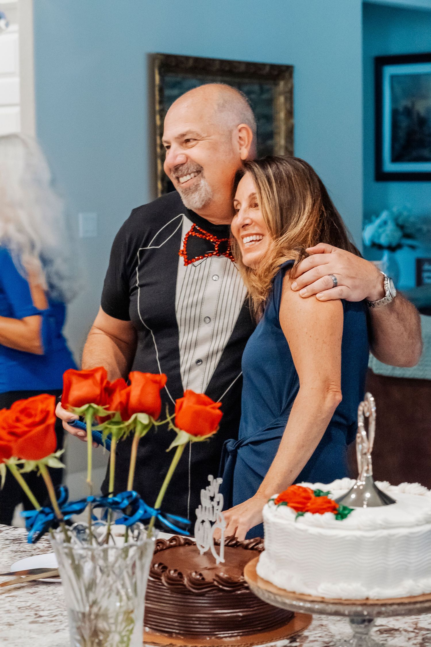 Couple embracing and smiling by a white wedding cake with red roses on a table at a celebration.