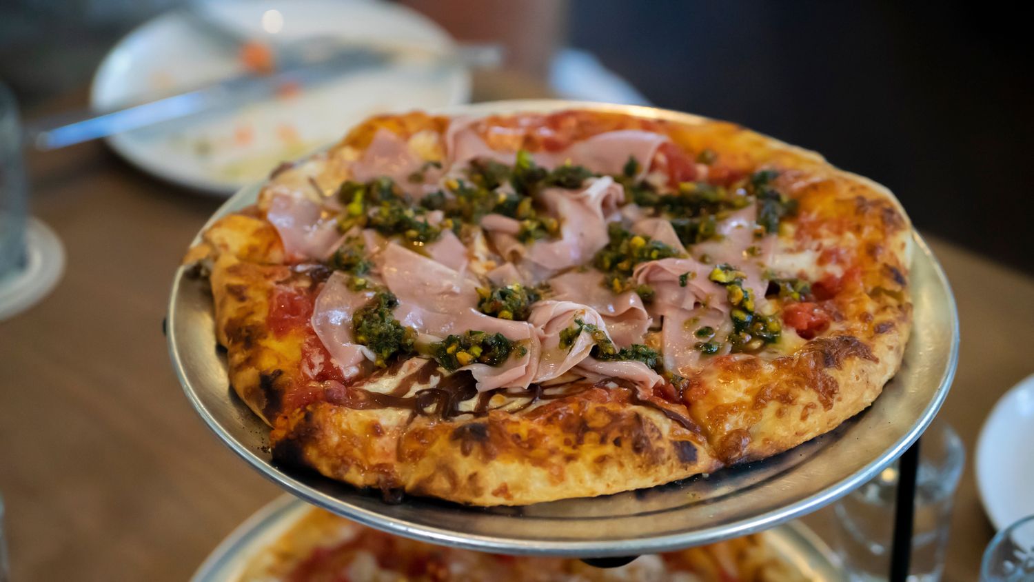 A Neapolitan-style pizza topped with ham, capers, and tomato sauce on a silver tray at a restaurant.