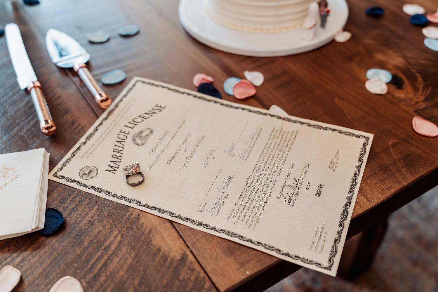 Marriage license on wooden table with wedding rings, cake, confetti, and cake server at a wedding reception.