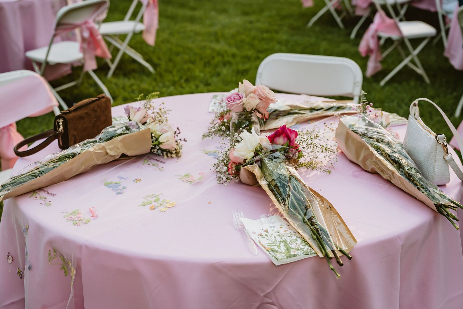 Pink round table with floral bouquets wrapped in greenery, set outdoors for an elegant garden wedding reception.