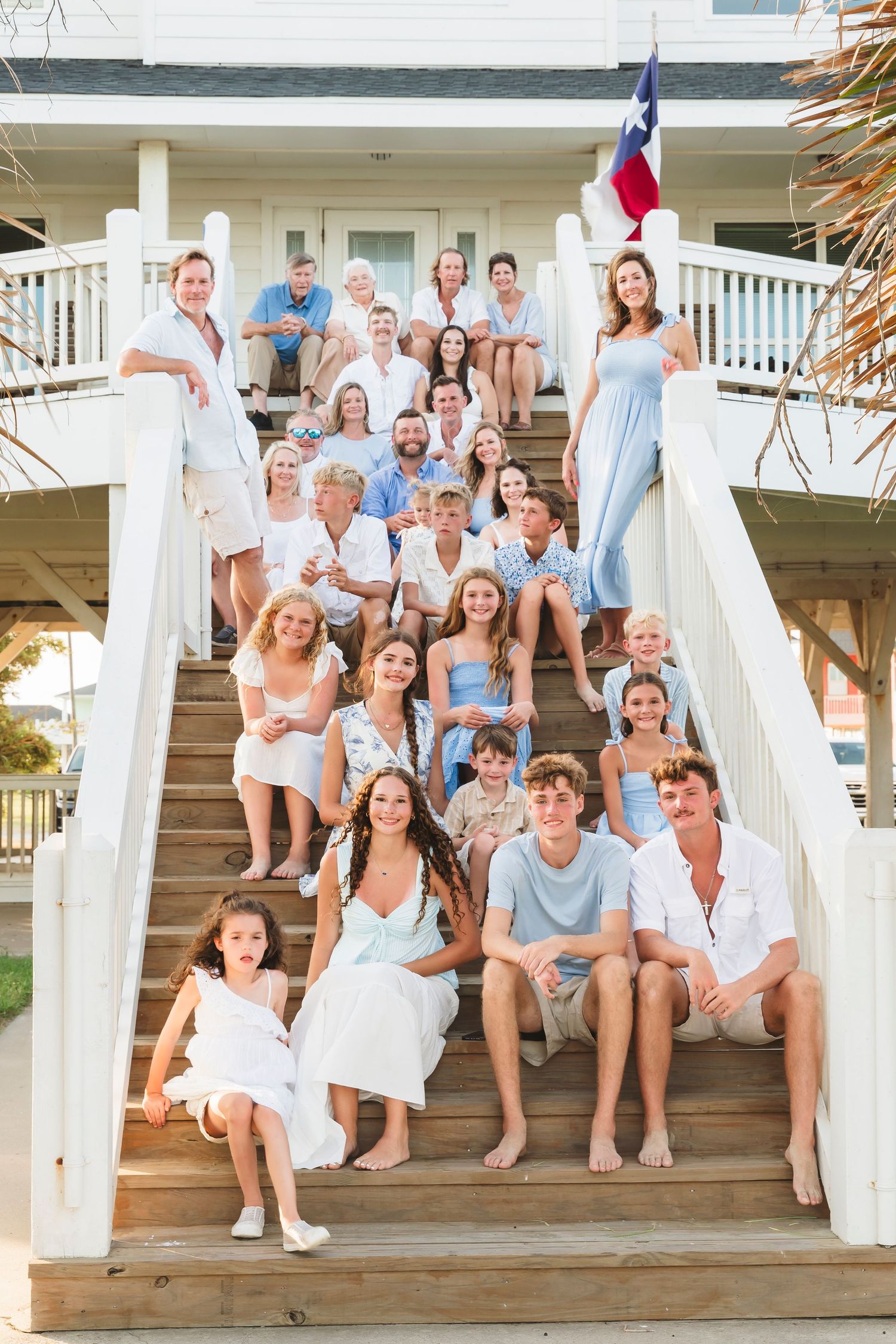 Large family group photo on beach house stairs, dressed in white and light blue, with Texas flag in background.