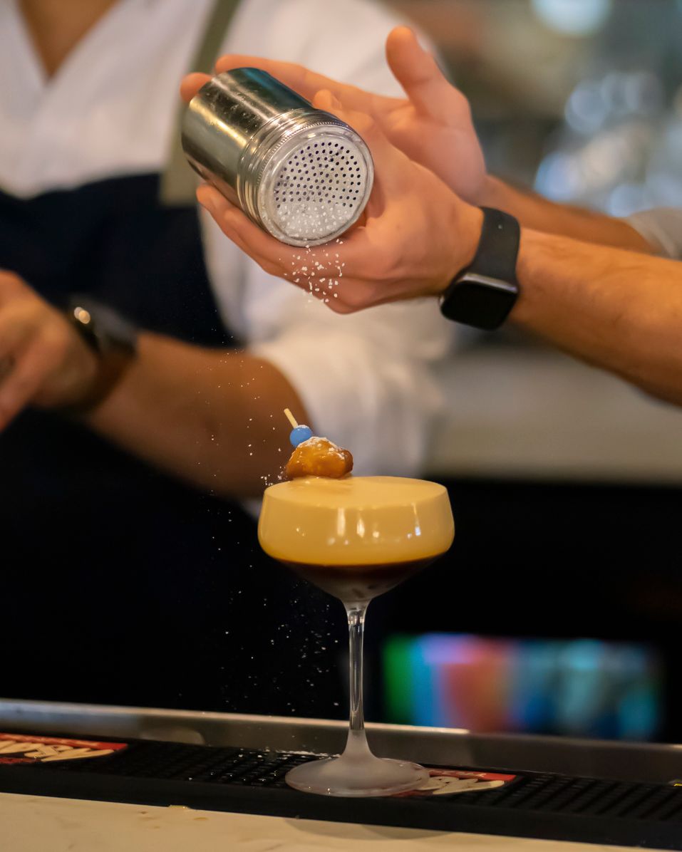 Bartender dusting spice over a frothy golden cocktail in a coupe glass at an upscale bar.
