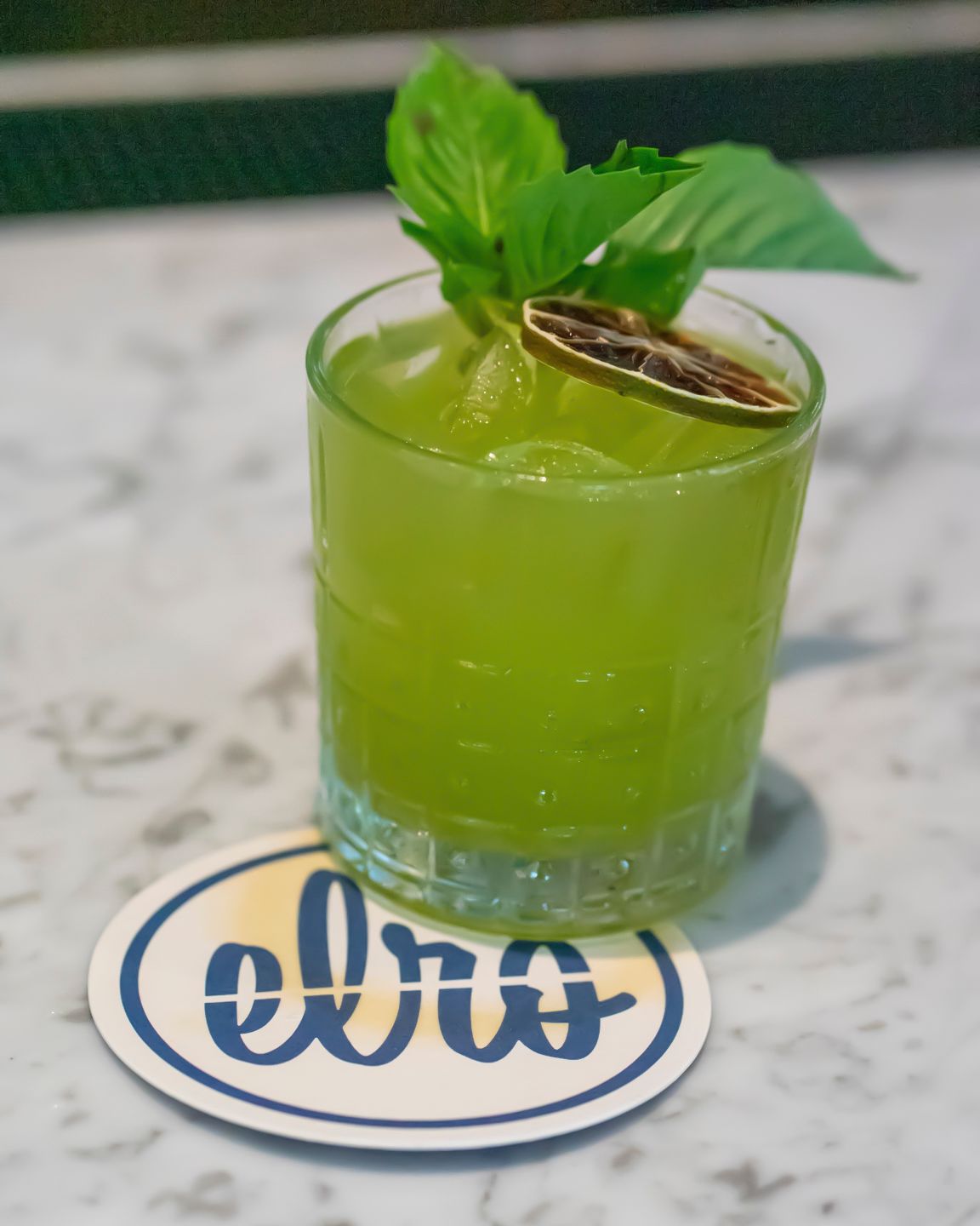 A vibrant green cocktail garnished with basil and a dried citrus slice on an Elro branded coaster.