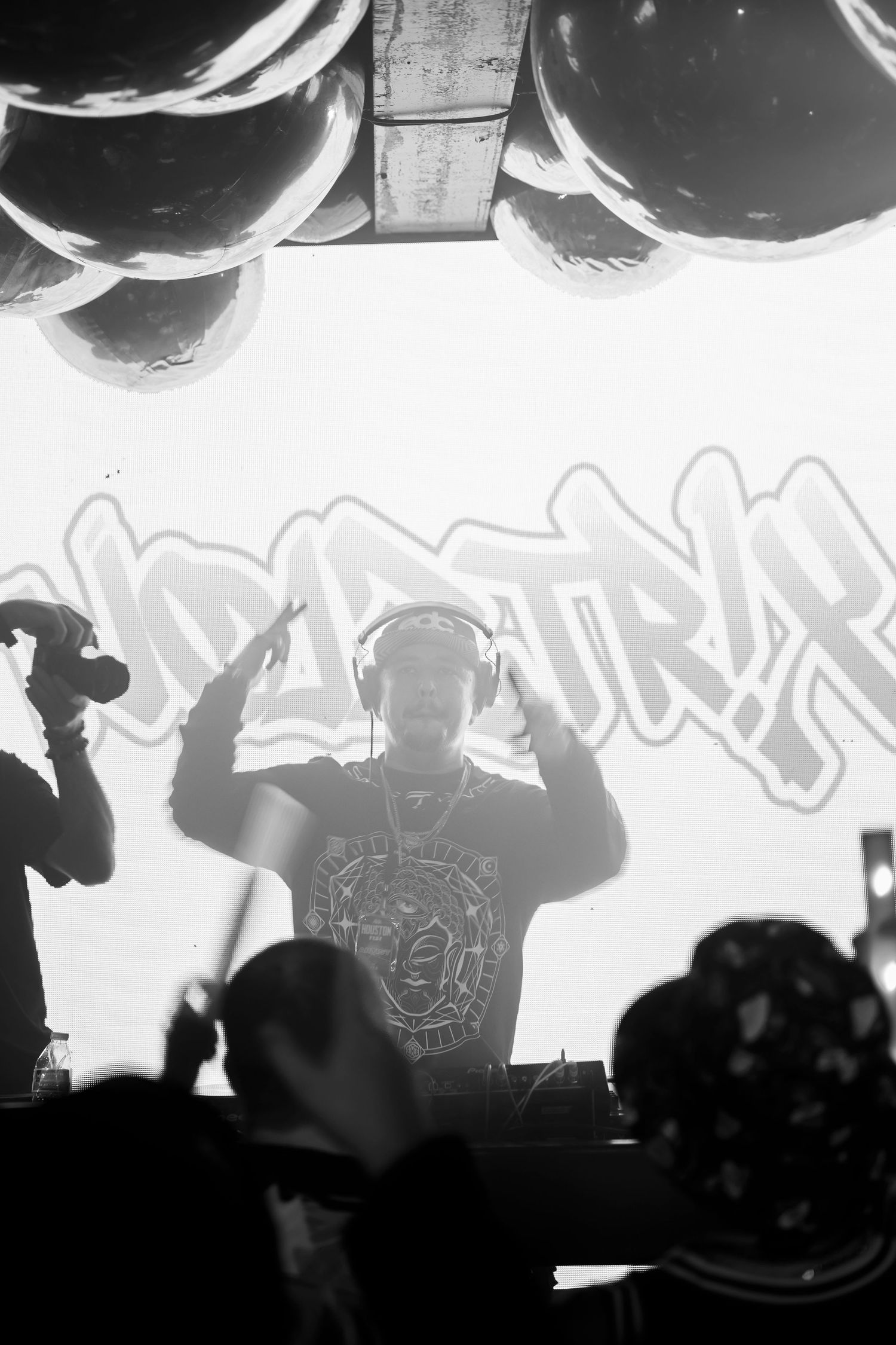 Black and white photo of a DJ performing on stage with headphones, graffiti backdrop, and balloons overhead.