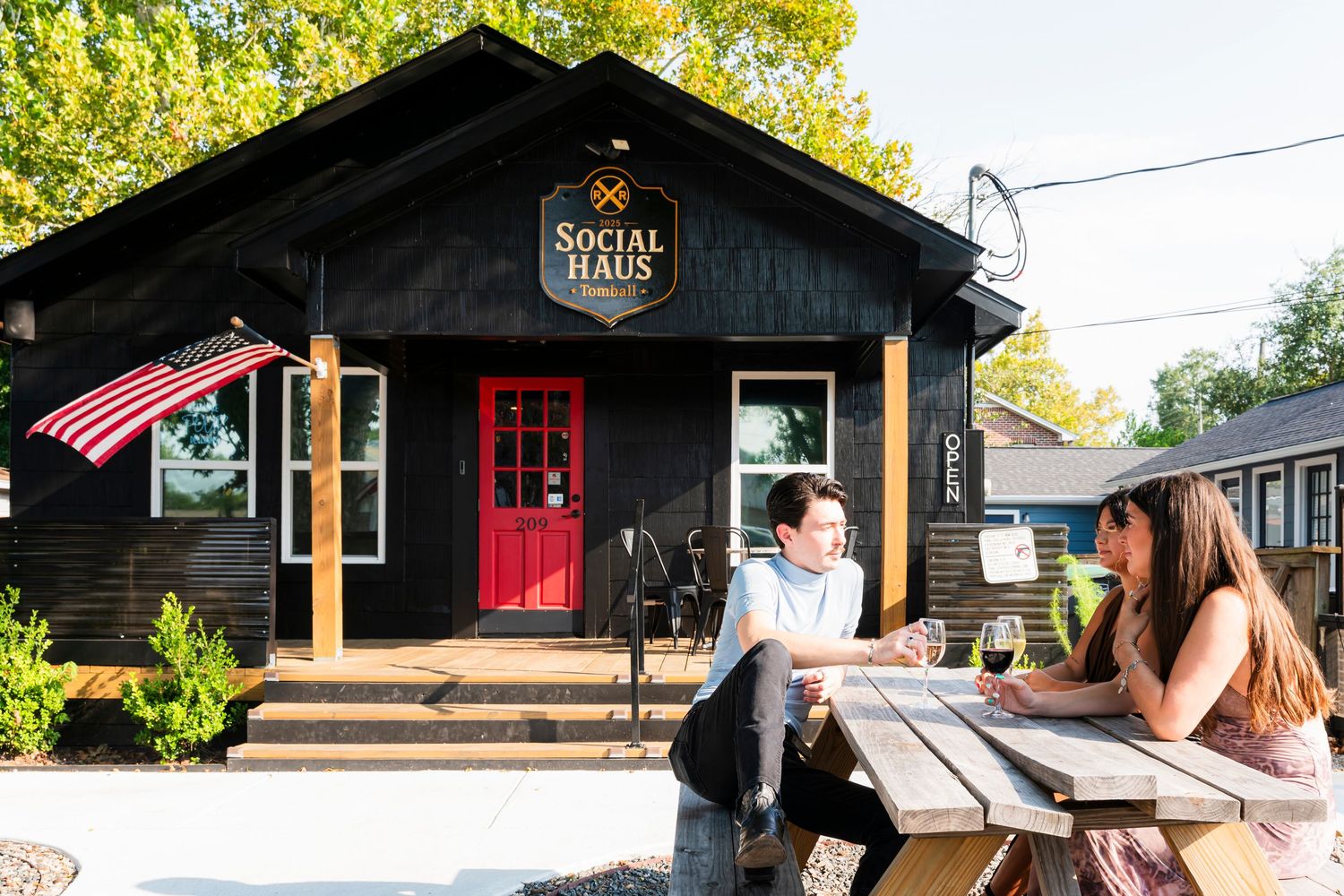 Two people enjoy drinks at an outdoor picnic table in front of Social Haus, a charming black wood-sided building.
