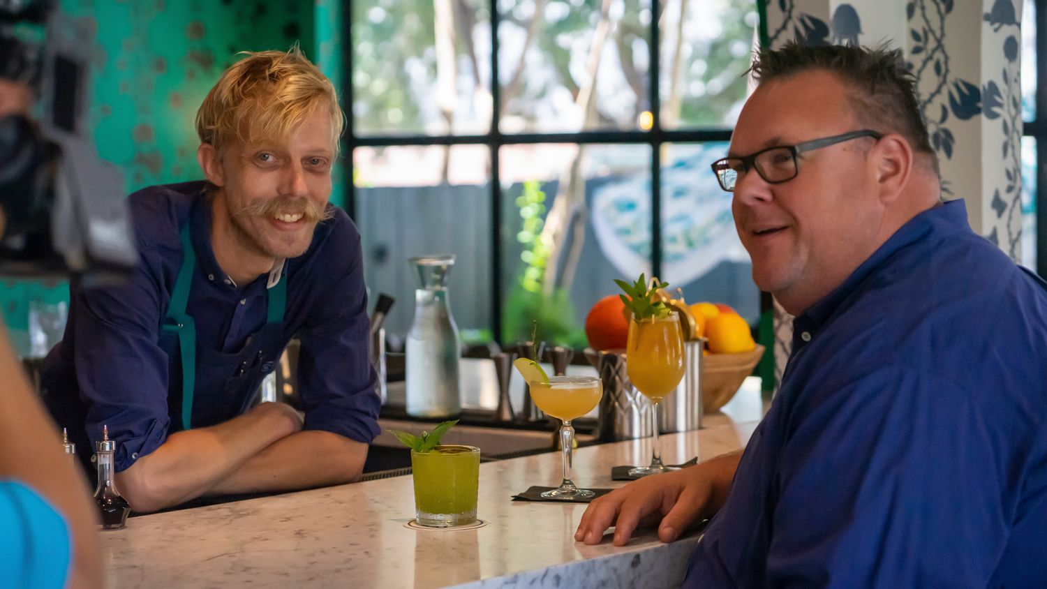Two men smiling and chatting at a bar counter with colorful cocktails and orange juice glasses in front of them.